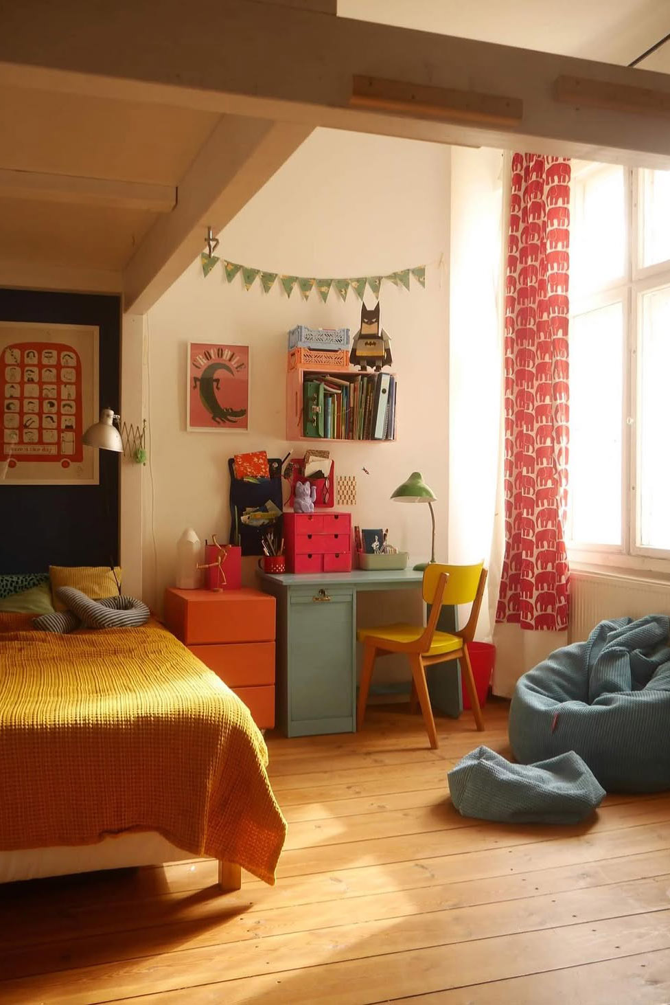Chambre d’enfant colorée avec lit jaune, bureau vert, chaise jaune, rideaux à motifs rouges et fauteuils en tissu bleu